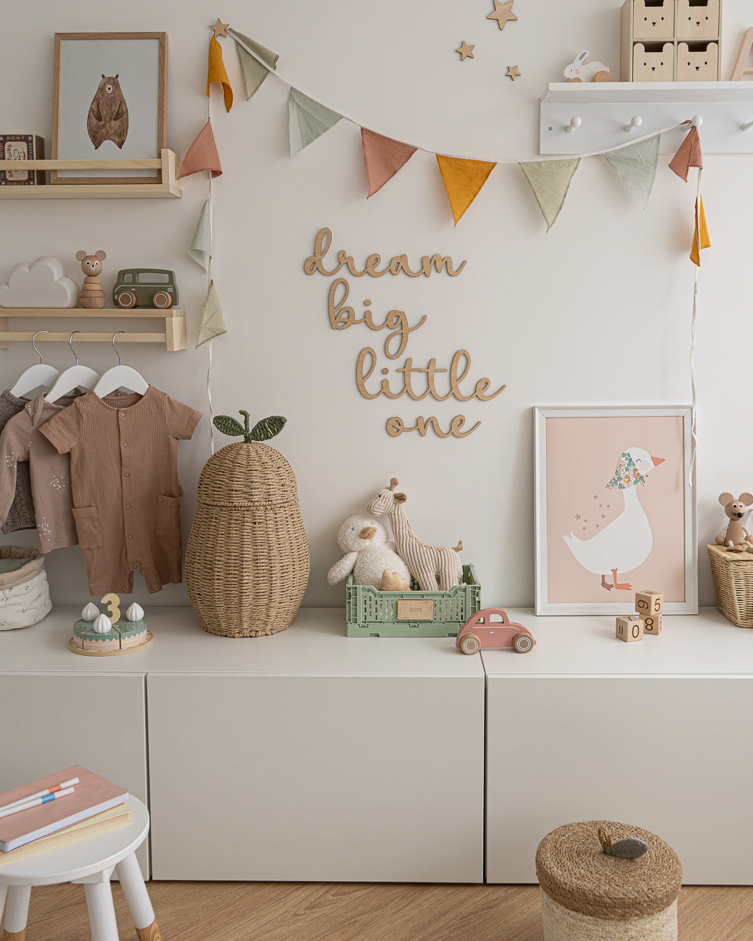 Wooden 'dream big little one' wall word sign in a script style font in a kids room displayed on a white wall, with pastel coloured accessories. 