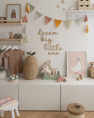 Wooden 'dream big little one' wall word sign in a script style font in a kids room displayed on a white wall, with pastel coloured accessories. 